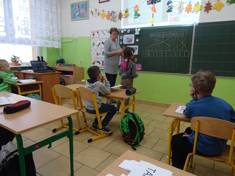 Klasa szkolna. Uczniowie siedzą w ławkach. Uczennica i nauczycielka stoją przy tablicy.