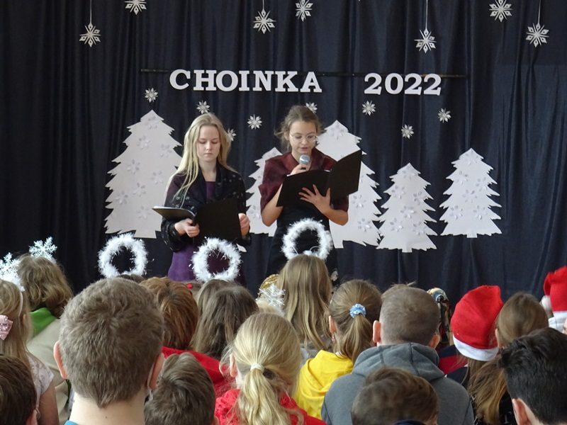 Korytarz szkolny. Na pierwszym planie dzieci siedzą na krzesłach. Przed nimi dwie uczennice z mikrofonem na tle napisu na ścianie: Choinka 2022.