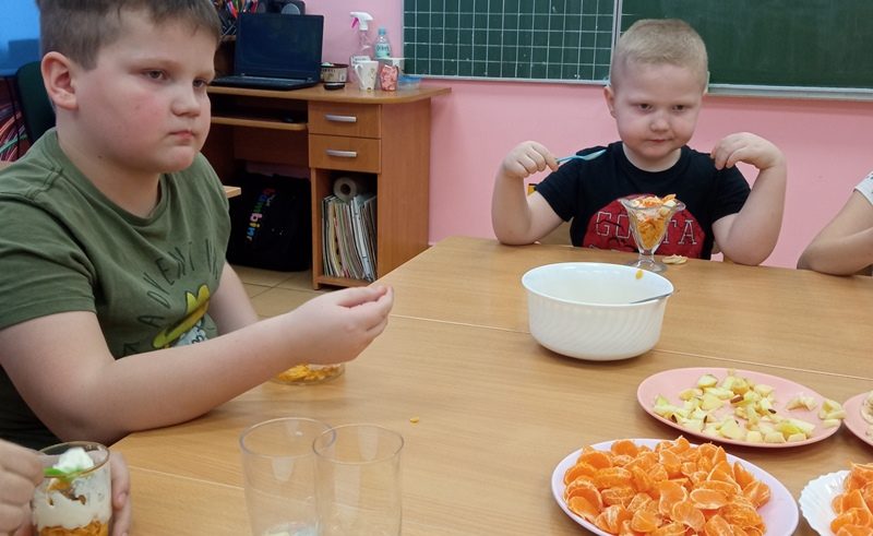 Klasa szkolna. Dzieci siedza przy stoliku i jedza deser. Na stoliku miska i talerze z pokrojonymi owocami.