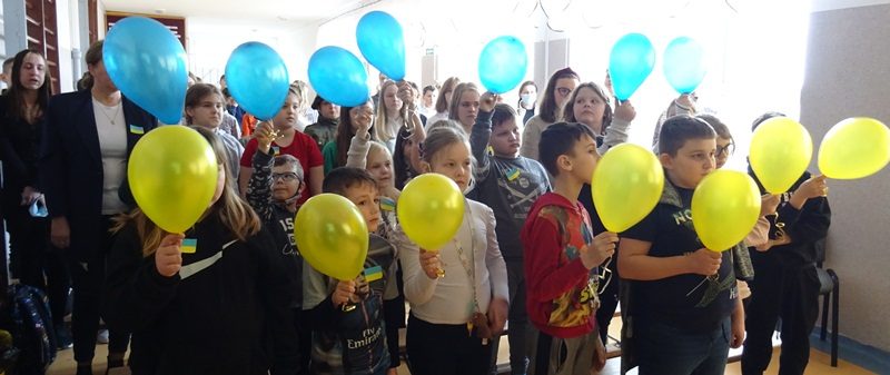 Korytarz szkolny. Wszyscy stoją. Uczniowie w pierwszym rzędzie trzymają żółte baloniki, a w drugim niebieskie.