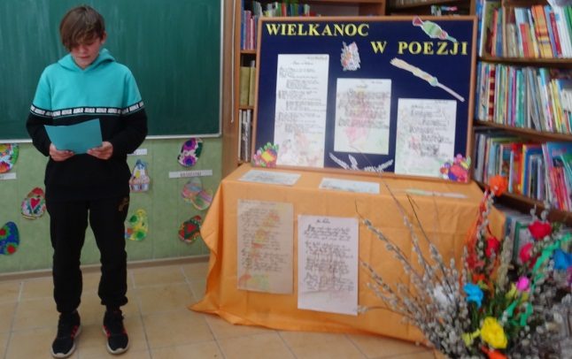 Biblioteka szkolna. Uczeń recytuje wiersz. Po prawej stronie dekoracja związana z  Wielkanocą.