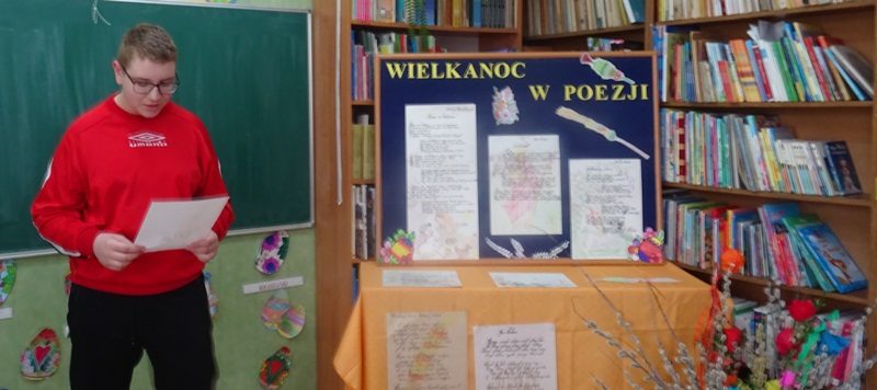 Biblioteka szkolna. Uczeń recytuje wiersz. Po prawej stronie dekoracja związana z  Wielkanocą.