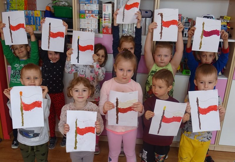 Sala przedszkolna. Dzieci stoją przy regale i trzymają rysunki przedstawiające flagę państwową.