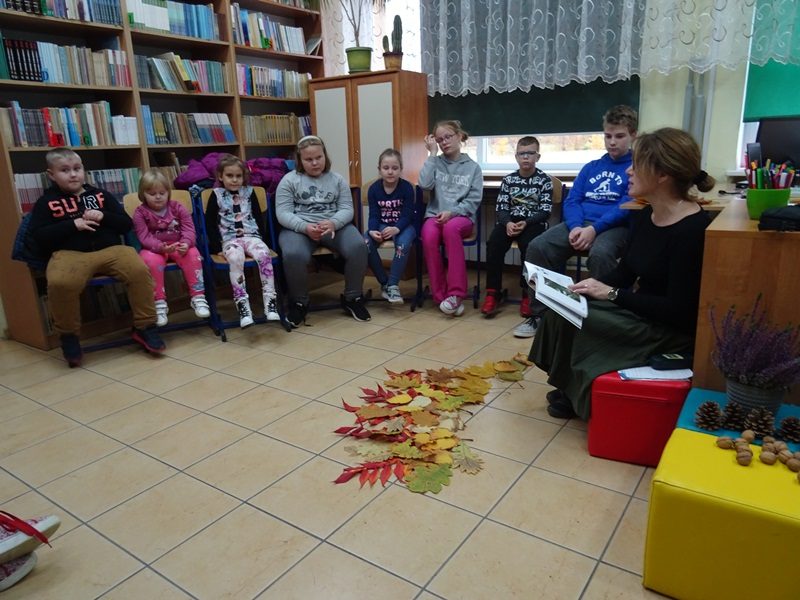 Biblioteka szkolna. Dzieci siedzą na krzesłach w półokręgu. Przed nimi siedzi nauczycielka i czyta opowiadanie. Przed uczestnikami spotkania na podłodze leżą jesienne liście.