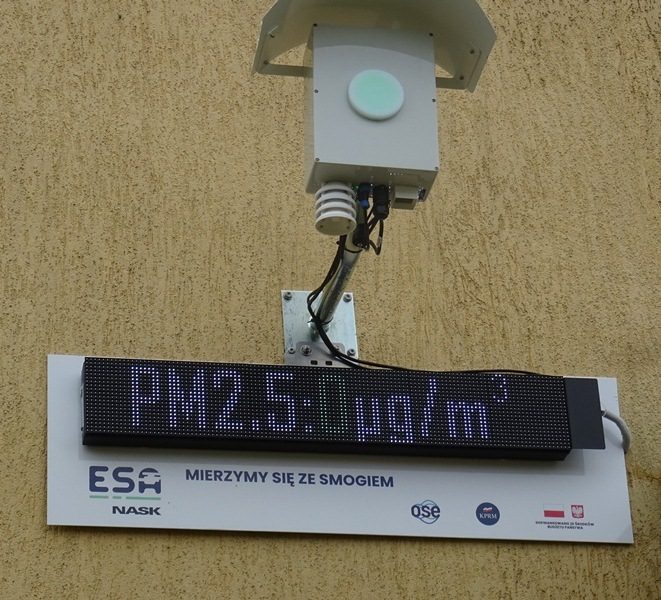 Budynek szkoły. Na ścianie zamontowany czujnik zewnętrzny smogu, który pokazuje wynik pomiaru: PM2.5: 0 ug/m3.