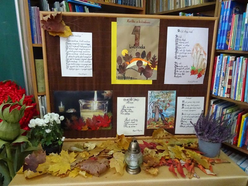 Biblioteka szkolna. Wystawka związana z 1 Listopada. Na stoliku stoją kwiaty w doniczkach i znicz oraz leżą suche, kolorowe liście.
