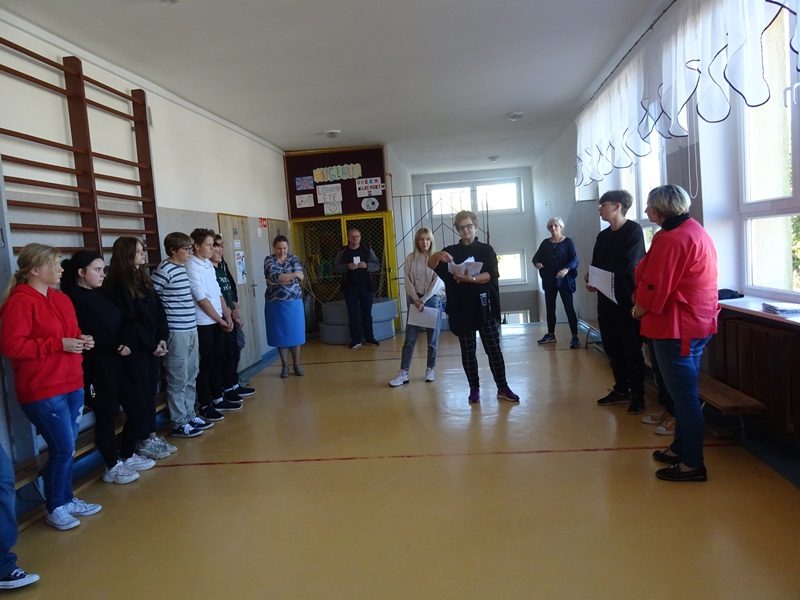 Korytarz szkolny. Uczniowie stoją przy ścianie, nauczyciele przy oknie i pośrodku korytarza. Nauczycielka prezentuje wyniki konkursu.