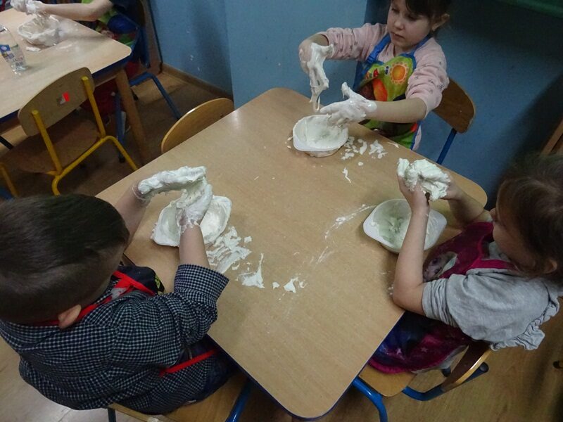 Sala przedszkolna. Dzieci siedzą przy stolikach i mieszają glutki w miseczkach.