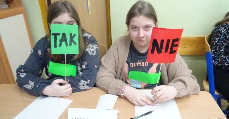 Sala lekcyjna. Dwie uczennice siedzą przy stoliku. W rękach trzymają napisy z wyrazami: Tak i Nie.
