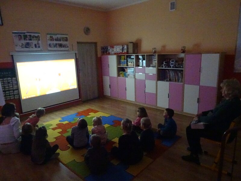 Sala przedszkolna. Dzieci siedzą na dywanie i oglądają film edukacyjny. Za nimi siedzi pielęgniarka szkolna.