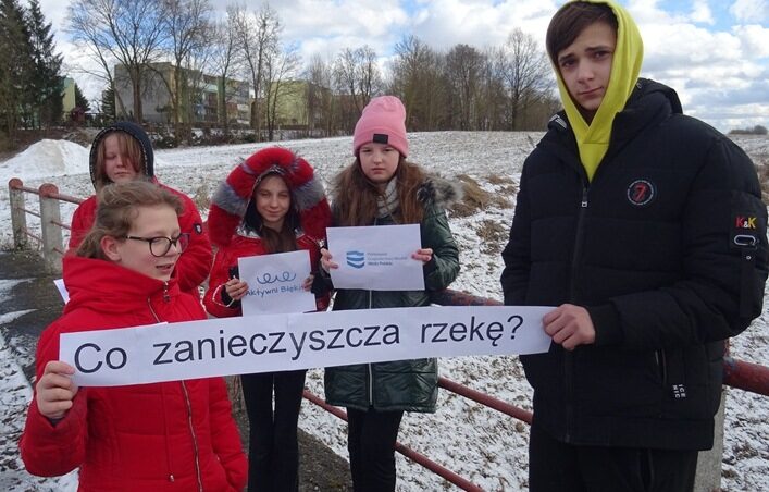 Nad rzeką Legą w Lenartach. Uczniowie stoją na moście i trzymają kartki z napisami: Aktywni Błękitni, Wody Polskie i Co zanieczyszcza rzekę?