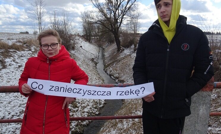 Nad rzeką Legą w Lenartach. Uczniowie stoją na moście i trzymają kartkę z napisem: Co zanieczyszcza rzekę?