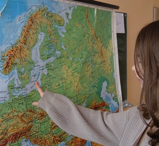 Sala lekcyjna. Uczennica stoi przy mapie i wskazuje na niej Morze Bałtyckie.