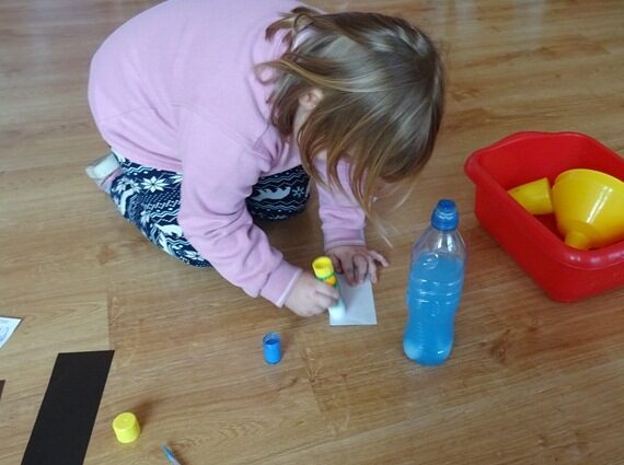 Sala przedszkolna. Dziewczynka smaruje klejem kartkę papieru. Przy niej stoi niebieska plastikowa butelka.