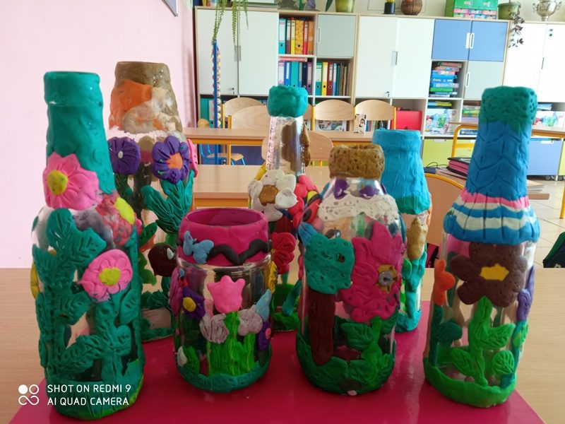 Klasa szkolna. Na stole prace uczniów: kolorowe wyklejanki na butelkach i słoikach.