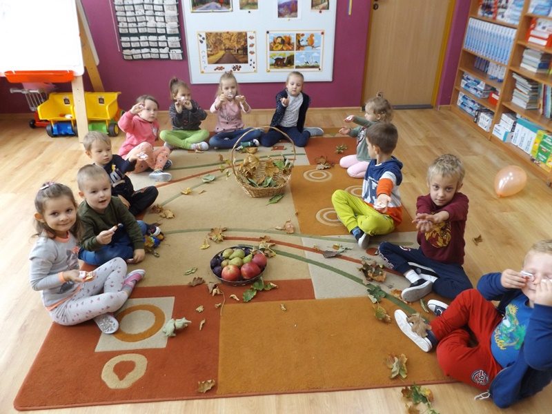 Sala przedszkolna. Na dywanie siedzą dzieci i bawia się liśćmi. Pośrodku dywanu stoi kosz z liśćmi i koszyk z owocami.