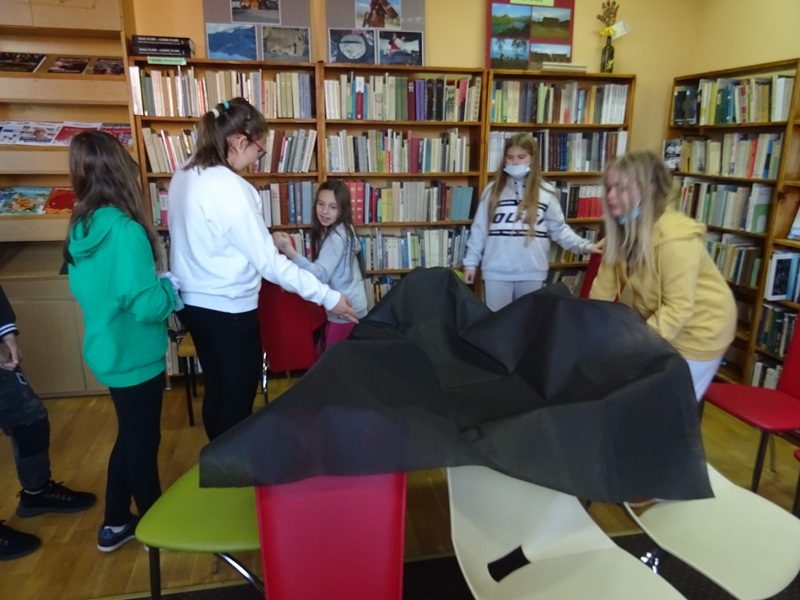 W bibliotece. Uczniowie chodzą dokoła krzeseł przykrytych czarną tkaniną.