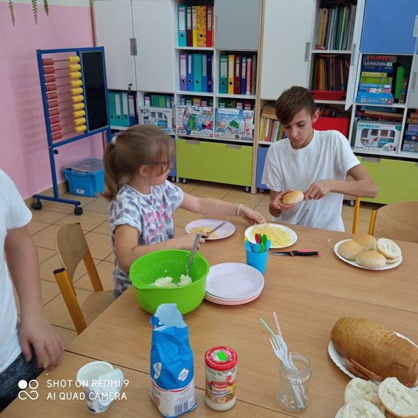 Klasa szkolna. Uczniowie przy stolikach przygotowują kanapki. Na stole leżą produkty spożywcze.