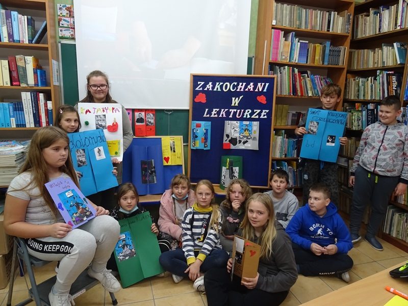 Biblioteka szkolna. Uczniowie stoją i siedzą przy tablicy i prezentują wykonane lapbooki.