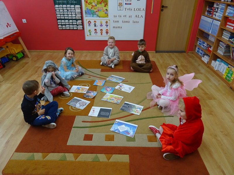 Sala przedszkolna. Dzieci w przebraniach siedzą na dywanie. Pośrodku dywanu leżą ilustracje przedstawiające fragmenty wybranych bajek.