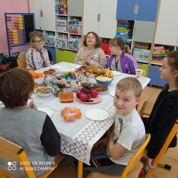 Sala lekcyjna. Pośrodku klasy stół nakryty białą serwetą. Na nim potrawy wigilijne. Przy stole siedzą dzieci.