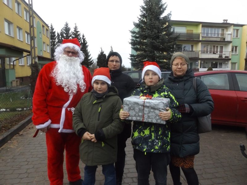 W Lenartach. Uczniowie, nauczycielki i Mikołaj stoją na chodniku przy blokach. Jeden z uczniów trzyma prezent.