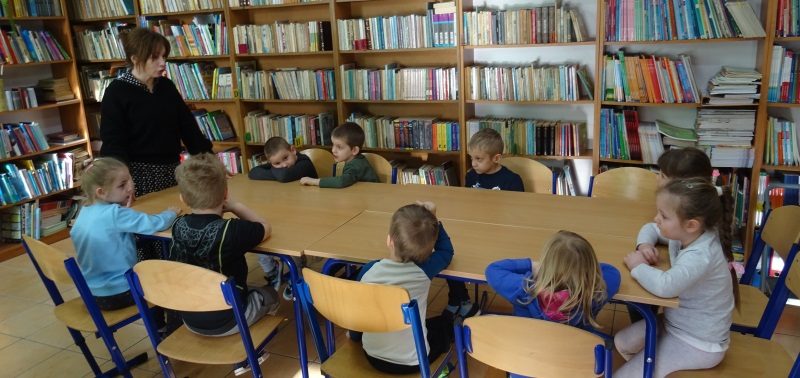 Biblioteka szkolna. Pośrodku połączone ze soba stoliki, przy których siedzą dzieci. Przed dziećmi stoi nauczycielka. Przy ścianach półki z książkami.