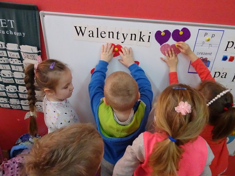 Klasa przedszkolna. Dzieci zaczepiają kolorowe serduszka na tablicy z napisem: Walentynki.