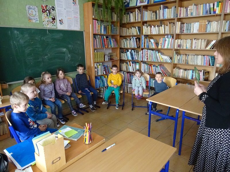 Biblioteka szkolna. Dzieci siedzą przy tablicy.  Przed nimi stoi nauczycielka.