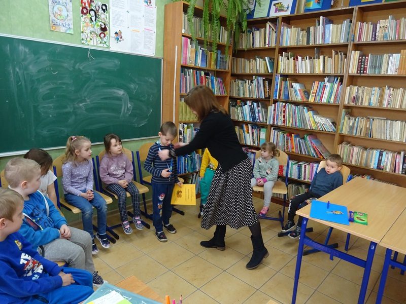 Biblioteka szkolna. Dzieci siedzą przy tablicy.  Nauczycielka wręcza dziecku nagrodę. 