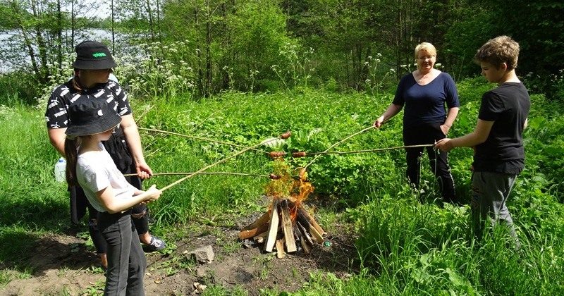 W lesie nad Jeziorem Czarnym w Judzikach. Pośrodku niewielkie ognisko, przy którym uczniowie pieką kiełbaski.