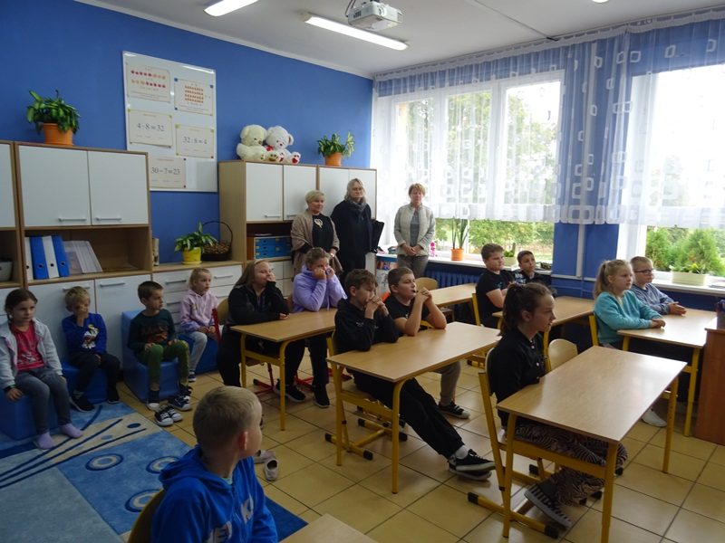 Sala lekcyjna. Uczniowie siedzą w ławkach i na pufach. Nauczyciele stoją przy szafie i oknie.