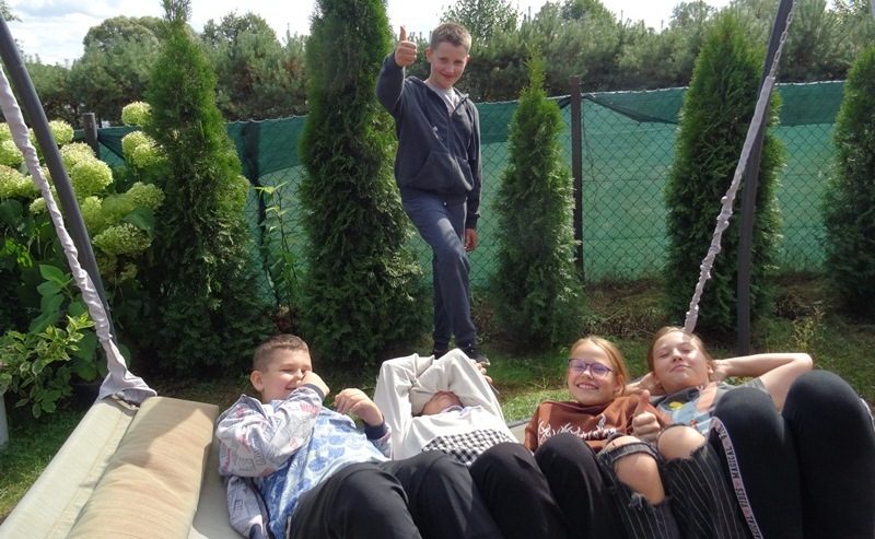 Małe Olecko. Uczniowie w ogrodzie. Leżą na huśtawce ogrodowej. Jeden uczeń stoi przed huśtawką. 