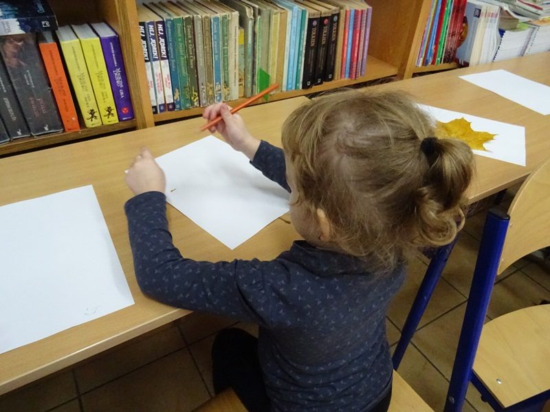 Biblioteka szkolna. dziewczynka siedzi przy stoliku i odrysowywuje liść.