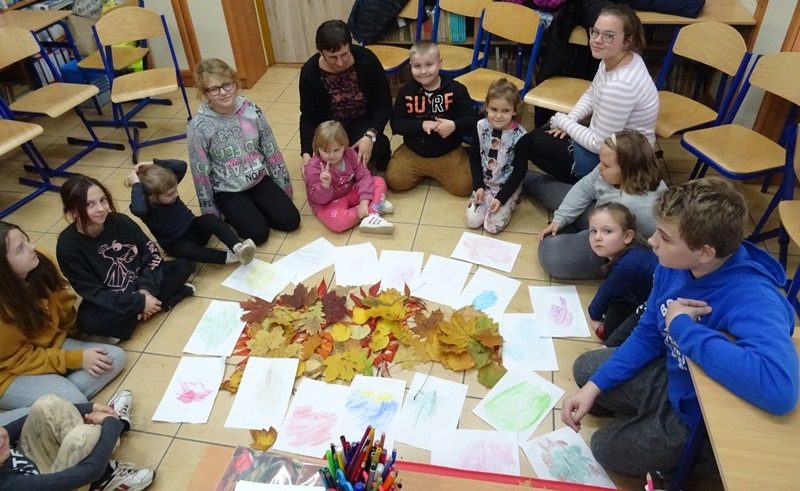 Biblioteka szkolna. Dzieci i nauczycielka siedzą na podłodze w półokręgu. Przed nimi na podłodze leżą jesienne liście i wykonane prace na białych kartkach papieru.