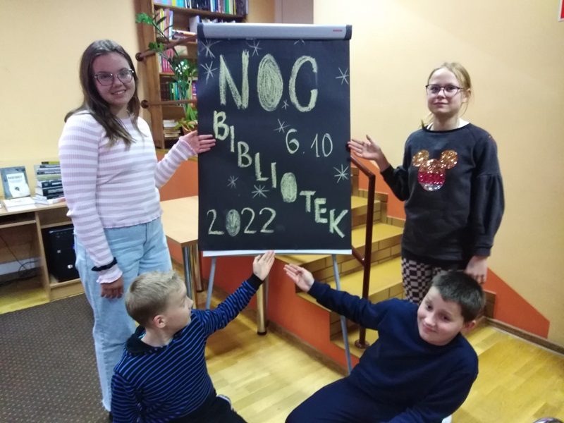 Biblioteka w Olecku. Uczniowie wskazują na napis umieszczony na tablicy: Noc Bibliotek, 6.10.2022.