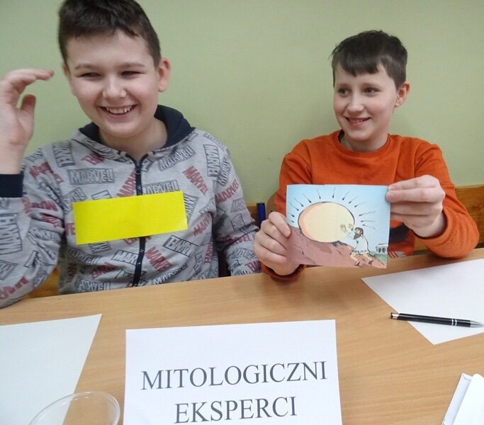 Sala lekcyjna. Dwóch uczniów siedzi przy stoliku, na którym jest napis: Mitologiczni eksperci. Jeden z uczniów trzyma rysunek przedstawiający mit.