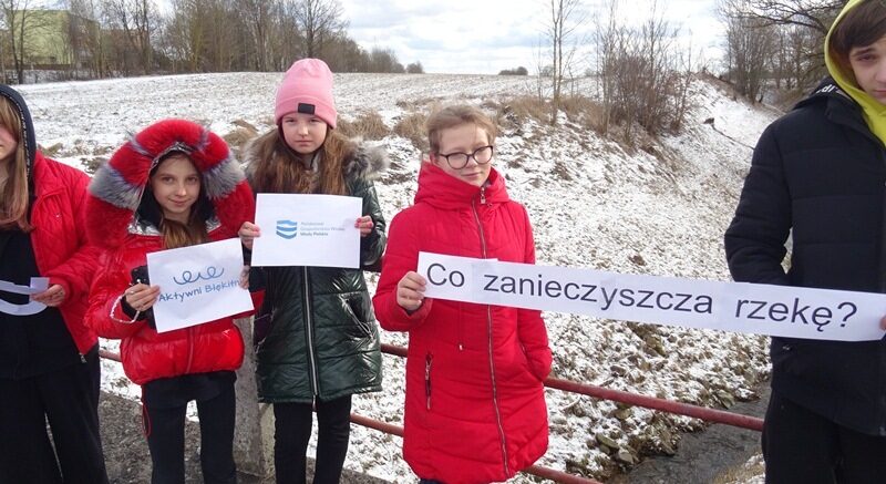 Nad rzeką Legą w Lenartach. Uczniowie stoją na moście i trzymają kartki z napisami: Aktywni Błękitni, Wody Polskie i Co zanieczyszcza rzekę?