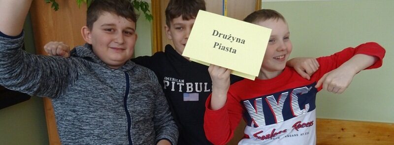 Sala lekcyjna. Uczniowie stoją obok siebie. Jeden z nich trzyma kartkę z napisem: Drużyna Piasta.
