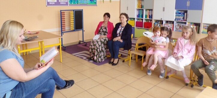 Sala dydaktyczna. Rodzic siedzi na krześle przed dziećmi i innymi rodzicami i czyta wiersz.