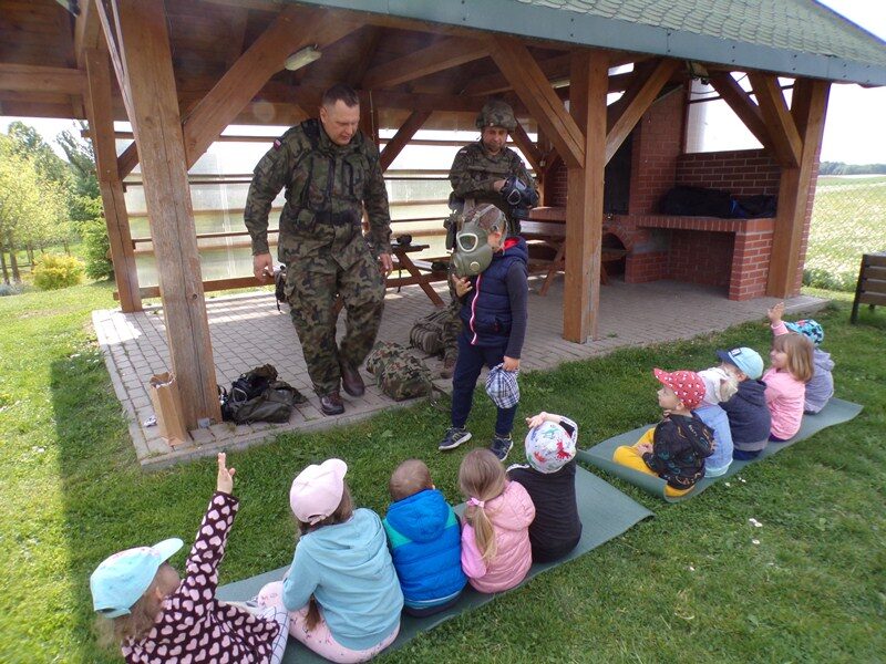 Posesja szkolna. Altanka. Dzieci siedzą na karimacie. Przed nimi stoją żołnierze. Dziecko ma nałożoną maskę przeciwgazową.