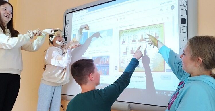Sala dydaktyczna. Uczniowie stoją przy tablicy interaktywnej i wskazują latarnie morskie na slajdzie.
