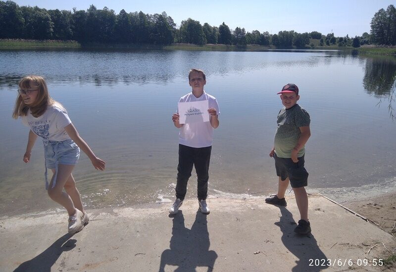 Nad Jeziorem Oleckim Wielkim. Uczniowie stoją na brzegu jeziora. Uczeń trzyma logo Aktywni Błękitni.