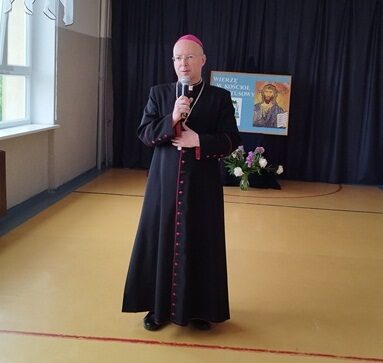 Korytarz szkolny. Ksiądz biskup stoi pośrodku i w ręku trzyma mikrofon.