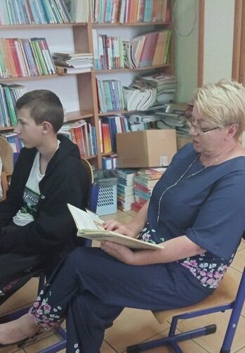 Biblioteka szkolna. Uczeń i nauczycielka siedzą na krzesłach. Nauczycielka trzyma w rękach ksiązkę.