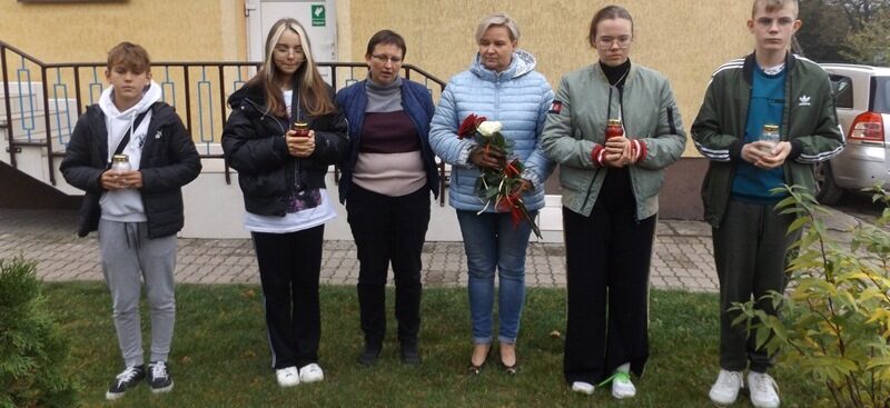 Posesja szkolna. Delegacja uczniów i dwie nauczycielki stoją obok siebie przed tablicą pamiątkową. Uczniowie trzymają znicze, a nauczycielka róże.