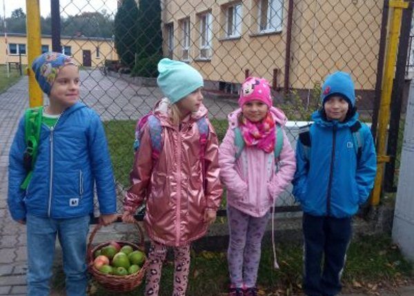 Posesja szkoły. Czterech uczniów stoi przed wejściem na posesję szkolną. Dwoje uczniów trzyma kosz z jabłkami.