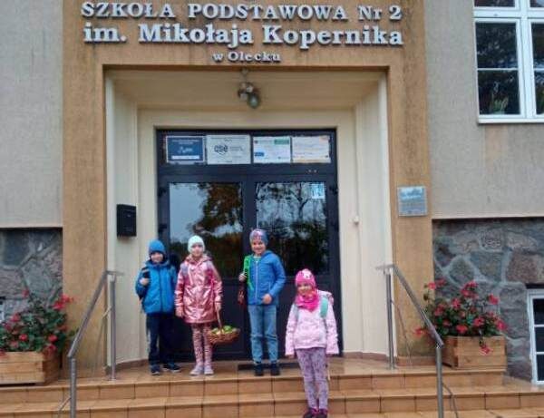 Posesja Szkoły Podstawowej nr 2 w Olecku. Czterech uczniów stoi przed wejściem do budynku.Uczennica trzyma kosz z jabłkami.