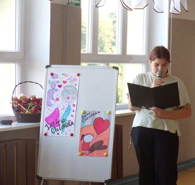 Korytarz szkolny. Uczennica trzyma mikrofon i czyta tekst. Obok niej na tablicy dwa rysunki z napisem Dzień Chłopaka.