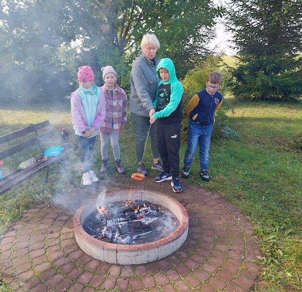 Posesja szkolna. Nauczycielka i uczniowie stoją przy ognisku. Uczeń trzyma na kijku dwie kiełbaski i piecze je przy ognisku.